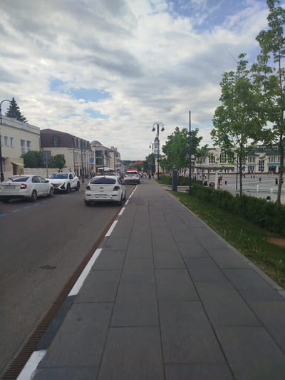 The image captures a street scene in Noginsk, Russia. A paved pedestrian walkway runs along the right side of the frame, bordered by a low hedge and young trees. To the left, a road contains parked cars, including a white sedan with the license plate P176PE799 and other vehicles with Russian license plates. Buildings line the street, with a mix of architectural styles. In the distance, people are seen walking and gathering in a square or plaza, and a building with an arch and signage is visible. The sky is overcast with patterned clouds.