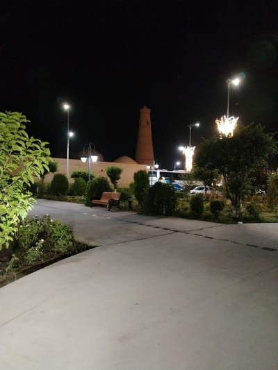 This image depicts a nighttime scene in a park or public square in Khiva, Uzbekistan. The foreground shows a paved walkway with a planter box containing greenery and a single white rose. A wooden bench with ornate metal legs sits on the walkway, inviting rest.

In the middle ground, a low, cream-colored wall runs horizontally, separating the paved area from a landscaped garden. Behind the wall, various bushes and trees are visible, some illuminated by streetlights. Two prominent lampposts with multiple globes cast a warm glow on the surroundings. Further back, a tall, cylindrical brick structure, possibly a minaret, stands against the dark night sky. Its tapered shape and the textured brickwork are distinct.

To the right, a large tree with a dense canopy is lit from below by a decorative light fixture shaped like a crown or a burst of flames, adding a festive ambiance. Several vehicles, including a white car and a bus with visible text on its side (partially obscured but appearing to be "229"), are parked or driving in the background, suggesting proximity to a road or parking area. The overall lighting suggests it is nighttime, and the air appears clear. The scene evokes a sense of calm and perhaps a tourist attraction open in the evening.