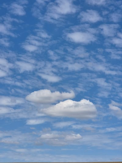 The image shows a vast expanse of blue sky filled with scattered white clouds. The clouds vary in size and density, with some appearing as wispy streaks and others as puffy, rounded formations. A thin, horizontal line of brown, likely the horizon, is visible at the very bottom of the image, suggesting a landscape below the sky. The scene is devoid of any people or specific man-made structures. The overall impression is one of an open, natural environment, possibly a rural or agricultural area given the hint of a cultivated land or field at the bottom. No specific details are discernible to pinpoint the location to the city of Mengkofen, Germany, but the scene is consistent with an outdoor setting in a temperate climate.