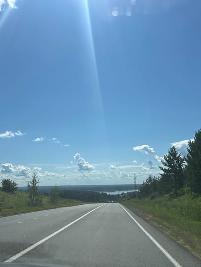 The image depicts a bright, sunny day with a clear blue sky. A paved road with white lane markings stretches into the distance, suggesting it is a highway or a major road. The road is bordered by green grassy hills and scattered trees on either side. In the background, there is a body of water, likely a lake or river, surrounded by a dense forest. A power line tower is visible in the mid-ground. The overall scene conveys a sense of open space and natural landscape, with no people or vehicles clearly visible in the immediate foreground or prominently within the scene. The sunlight creates a lens flare effect in the upper left portion of the image. The location is identified as Nechkino, Russia.