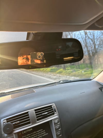 The image depicts the interior of a vehicle, viewed from the driver's perspective, located in Musachevo, Bulgaria. The central focus is the rearview mirror, reflecting a person's face and hand. The person, with facial hair including a mustache and beard, has a black nasal strip applied across their nose and is holding a dark-colored smartphone with multiple cameras, actively taking a picture of their reflection.

The dashboard, colored grey and black, is visible in the foreground, featuring air vents, an infotainment system with a "CAUTION" message displayed on its screen, and buttons labeled "MAP," "DEST," and "MENU." A circular Hama brand phone mount is affixed to the dashboard near the infotainment screen. The passenger-side dashboard section has "SRS AIRBAG" embossed. The windshield, showing some dust, reveals a paved road directly ahead of the vehicle. Beyond the road, green ground vegetation and deciduous trees with bare branches are visible under a clear, light blue sky. Strong, low-angle sunlight enters from the left, creating lens flare and bright reflections within the vehicle interior.