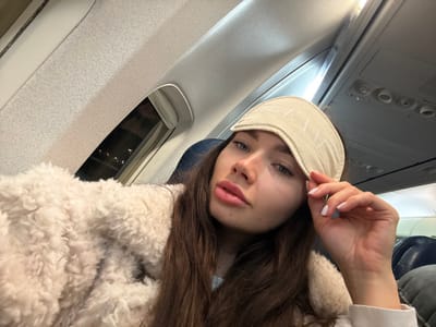 A woman on an airplane poses for a selfie. She is wearing a fuzzy cream-colored coat and has long brown hair. She holds a sleeping mask in her hand, partially covering her forehead. The airplane window shows a dark night sky with distant city lights. The airplane's interior, including the ceiling, seat, and side panel, is visible, adding context to the scene. The overall image has a slightly rotated perspective.