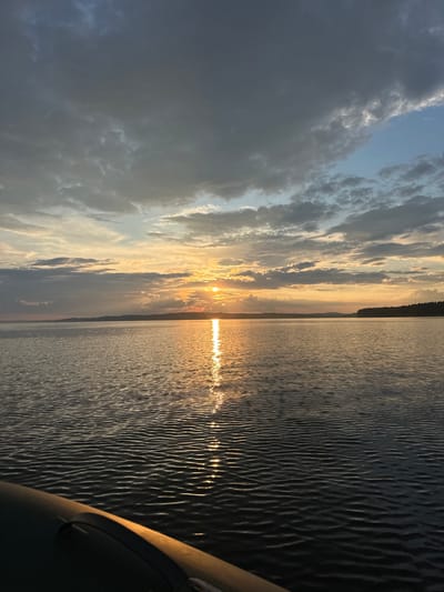 The image captures a serene sunset over a body of water, likely a lake or river, near Sosnovka, Russia. The sky is a dramatic canvas of gray and white clouds, with the setting sun casting a warm, golden glow in the center, creating a shimmering reflection on the water's surface. The horizon is marked by a dark, tree-lined shore. In the foreground, the edge of a dark inflatable boat is visible, suggesting the photograph was taken from its perspective. The water is calm, with small ripples distorting the reflection of the sun. The overall atmosphere is tranquil and picturesque, indicative of a peaceful evening by the water.