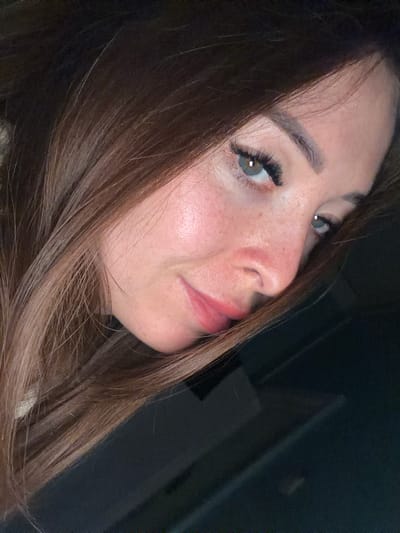 This media file shows a close-up, angled portrait of a woman's face. She has long, brown hair that partially obscures her face. Her eyes are light green and appear to have false eyelashes applied. She has a light complexion with visible freckles on her cheeks and nose. Her lips are painted with a glossy pink lipstick, and her mouth is slightly open, giving the impression that she might be speaking or about to speak. The lighting suggests it could be indoors, possibly in the evening or in a dimly lit environment, as the background is dark and out of focus. There are no discernible objects, text, or specific location cues within the frame that would provide further context about the scene or activity. The overall mood is somewhat intimate due to the close-up framing.