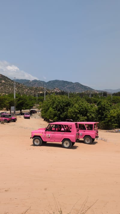The image depicts a pink Jeep-like vehicle parked on a sandy surface, likely a road or open area. In the background, there are mountains, lush green trees, and utility poles. Additional pink Jeep-like vehicles can be seen farther down the road. The location is likely Phuoc Dinh, Vietnam, based on the prompt.