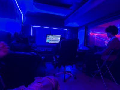 A dimly lit recording studio bathed in blue light showcases two people working. One sits in a rolling chair facing a computer screen displaying audio editing software. The other person, wearing glasses and a cardigan, sits facing away in a folding chair, gazing through a large window. Neon lights are reflected in the glass of the window. A third person is partially visible at the bottom. The studio walls are lined with neon lighting.