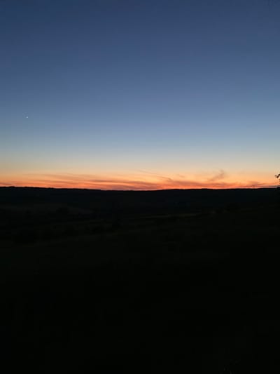 The image captures a twilight scene over a rural landscape, potentially in or near Negrești, Moldova. The sky transitions from a deep blue at the top to a soft gradient of lighter blue and then a vibrant band of orange and pink hues near the horizon, indicating the last moments of sunset. Wispy clouds are visible within the colored band, catching the fading light.
Below the sky, a dark silhouette of rolling hills and trees forms the horizon line. The foreground is almost entirely black, suggesting it is a very dark field or wooded area, heavily underexposed. A single bright point, possibly a star or planet, is visible in the upper left portion of the sky, and another fainter one is near the horizon on the right. There are no people or distinct objects visible in the foreground or midground due to the low light conditions. The overall impression is one of tranquility and natural beauty at dusk.