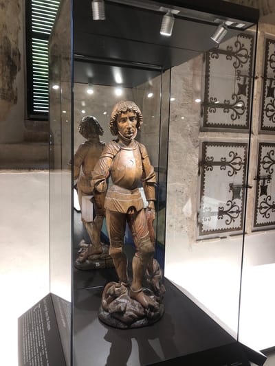 The image shows a wooden sculpture of Saint George slaying a dragon, displayed in a glass case. The sculpture depicts a knight in armor standing over a dragon. The case is in front of a stone wall with decorated panels on it. The sculpture is described by a plaque next to it, detailing the sculpture's origins and history. The location, according to the text, is the Chillion chapel of Veytaux, Switzerland.