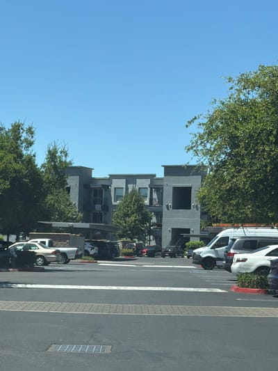 The image shows a parking lot in front of a modern, multi-story apartment building with a gray facade, visible under a clear blue sky. Several vehicles are parked in the lot, including a white van, a dark SUV, and a white sedan. Some greenery, including trees and bushes, is interspersed with the buildings and parking lot. The scene appears to be in a developed urban or suburban area, consistent with a location in Elk Grove, United States of America.
