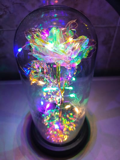 A holographic rose, illuminated by a string of colorful LED lights, is encased in a clear glass dome. The rose, made of iridescent material, catches and reflects the light, creating a spectrum of colors. The background is a blurred purple wall. The image does not provide any information about the city of Лосева, Belarus, or any people or actions.