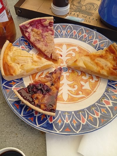 Four slices of pie are arranged on a decorative plate with a blue and orange pattern. One slice appears to be a berry pie, with visible blueberries and a dark, glossy filling. Another slice is an apple pie with golden-brown crust and light-colored fruit filling. A third slice is a fruit crumble with a pinkish-red filling and a crumb topping. The fourth slice is also a fruit pie, possibly apple, with a flaky crust and visible fruit. The plate is on a speckled gray surface, possibly a table. To the left of the plate is a glass bottle with a red label and a liquid inside. In the background, a tray with a rooster design is visible, along with a white cup and a blue thermos. The context provided is that this scene is located in Murten, Switzerland.