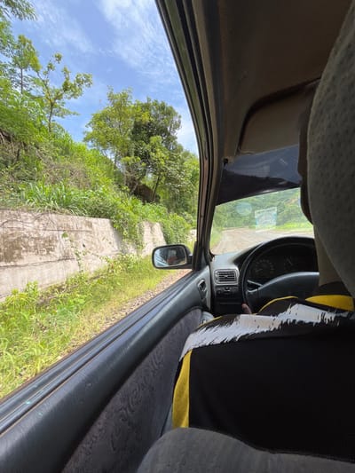 The image shows a first-person view from the passenger seat of a moving car on a dirt road, likely in Goje, Nigeria. Lush green vegetation, including tall grasses and trees, borders the road on the left. A concrete retaining wall runs alongside the road, with more greenery growing over and around it. The road itself is unpaved and appears to be bumpy, with evidence of tire tracks. In the distance, the road curves around a densely vegetated hillside. Inside the car, the dashboard, steering wheel, and part of the driver's shoulder and head are visible. A black and yellow patterned fabric covers the passenger seat. The sky is blue with scattered white clouds.