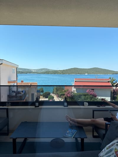 The image shows a view from a balcony overlooking a body of water in Grad Šibenik, Croatia. In the foreground, a dark coffee table with a book on it is visible, along with a person sitting in a chair with their legs extended. A white mug and a pair of sunglasses are on the balcony railing. The middle ground features houses with red tiled roofs and lush greenery. The background displays a calm blue sea with hills covered in trees on the horizon under a clear blue sky.