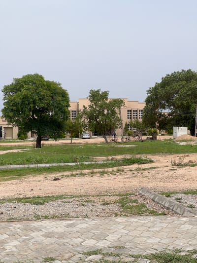 This is a vertical, outdoor, eye-level shot of a building and its surroundings in Yali, Nigeria. The scene is dominated by a light beige, multi-story building with many windows. The building appears to be an institutional or educational facility. In front of the building, a large, mature tree with lush green foliage stands prominently. Smaller trees and shrubs are also scattered around the building.
Adjacent to the building and the trees is a dirt path, interspersed with patches of green grass and gravel. Further to the right, a paved walkway made of light-colored, diamond-shaped interlocking stones runs along the edge of the dirt path. The sky is overcast and a light, pale blue, suggesting a diffused light, possibly indicating early morning or late afternoon, or a cloudy day.
There are at least two vehicles visible near the building, a silver SUV and a black car, parked on what appears to be a driveway or access road. The overall impression is one of a quiet, perhaps academic or administrative, setting during daylight hours. No people are clearly visible in the scene, though some shadows might suggest their presence or recent activity. The environment appears dry, with sparse vegetation in some areas, characteristic of a warm climate. There is no discernible text visible in the image.