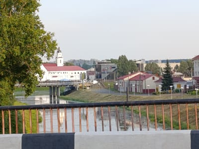 The image depicts a scene in the city of Orsha, Belarus, featuring a river and buildings. In the foreground, a dark metal railing with vertical copper-colored bars separates the viewer from the river. The river's surface reflects the sky and some of the surrounding structures.
On the far bank of the river, a white building with a red roof and a clock tower is visible. This structure appears to be a prominent landmark, possibly a church or a government building. To the right of this building, a bridge spans the river. Further along the riverbank, several other buildings with red roofs are present, including a white structure with a dark roof and windows, and a smaller white building with a red roof and multiple windows. A tall, dark green evergreen tree stands near these buildings. In the distance, a cityscape of residential buildings with light-colored facades and darker roofs is visible under a hazy sky.
The environment is characterized by a calm river, a green grassy bank, and a mix of architectural styles in the buildings. The sky is a pale, overcast blue. The overall impression is of a moderately urban landscape in daylight. No people are clearly visible in the image. The image is taken from a slightly elevated position, looking across the river towards the city.
