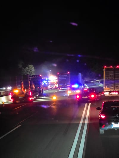 The image depicts a nighttime scene on a highway, likely in the vicinity of Seligenstadt, Germany, due to the license plate visible on a car. Several vehicles are present, including large trucks and a police car with flashing blue lights. One truck has a sign that reads "There's more to Prime. A truckload more." The road is well-lit by the vehicles' headlights and taillights, as well as by streetlights in the distance. The overall impression is one of a minor incident or a traffic stop. There is some light fog or mist in the air, causing reflections from the lights. The environment appears to be a multi-lane highway.