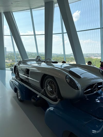 The image shows a silver Mercedes-Benz 300 SLR Uhlenhaut Coupé on display in the Mercedes-Benz Museum in Stuttgart, Germany. The car is positioned on a platform above a blue classic Mercedes-Benz. The museum's architecture features large, slanted windows that offer a view of the city and a partly cloudy sky. Several people are visible in the background, looking at the car. The floor of the museum is a polished gray surface.