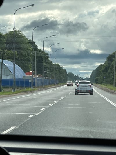 The image shows a road in Bol'shoe Teshkovo, Russia, with a cloudy sky overhead. On the left side of the road, there is a blue building with a curved metal roof, possibly a warehouse or industrial building. A row of streetlights is visible along the road. Several cars are driving on the road, including a white SUV towing a trailer in the foreground and a red car further ahead. The road is paved and has lane markings. Trees line both sides of the road, with denser foliage on the right. The overall scene depicts a typical suburban or rural highway environment.