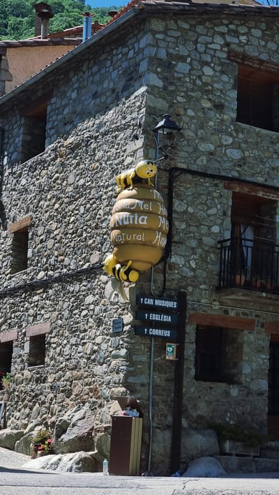 The image shows a street corner in the town of Setcases, Spain. A stone building with wooden window frames and a red tile roof is the main focus. A large, yellow, beehive-shaped sign with cartoon bees on it hangs from the corner of the building. Text on the sign reads "Nuria Mel Natural Hon". Several directional signs are posted on a wooden pole next to the building. One sign points upwards and reads "CAN MUSIQUES", another points upwards and reads "ESGLÉSIA", and a third points upwards and reads "CORREUS". Below these, a sign with a heart symbol and the letters "DEA" is visible, along with arrows indicating directions. A brown trash receptacle stands near the base of the pole. In the foreground, a paved road is visible, with some rocks and a small plant at the base of the building. The background features a lush green hillside with trees. The sky is clear and blue.