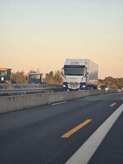 This image depicts a highway scene at dusk, possibly in Bischofsheim, Germany, with a focus on a white and blue semi-truck from the company "ETTIDE" on the right side of the road. The truck has the license plate "NX-7896". To the left of the semi-truck, there are two construction vehicles visible. The foreground shows a dark asphalt road with white and yellow lane markings. The sky transitions from a pale blue at the top to a warm orange and pink gradient towards the horizon. A small airplane is visible in the upper portion of the sky. The road is bordered by a concrete barrier.