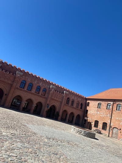 An upward-angled outdoor shot captures the architectural details of a historical building, likely a castle or fortress, under a clear blue sky in Guryevsk, Russia. The scene primarily features two sections of a red-brick structure and a cobblestone courtyard.
On the right side of the image, a building with a reddish-orange tiled roof is visible, featuring multiple rectangular windows with dark frames, some having arched tops. Below this, and extending across the middle of the frame, is a long, prominent brick wall adorned with numerous arched windows and deeper arched recesses or entrances, suggesting a ground level or a series of cloisters. This wall also displays decorative crenellations or battlements along its top edge. The red bricks vary in tone, contributing to the aged appearance of the structure.
The ground is paved with irregular cobblestones, showing various shades of gray and brown, indicating an old, well-worn courtyard. A low stone wall or planter is partially visible on the right side of the courtyard. The sky above is a uniform, vibrant blue, signifying a bright, clear, and sunny day, likely during late morning or early afternoon. No people or discernible text are visible within the frame, creating a quiet and expansive view of the historic architecture.