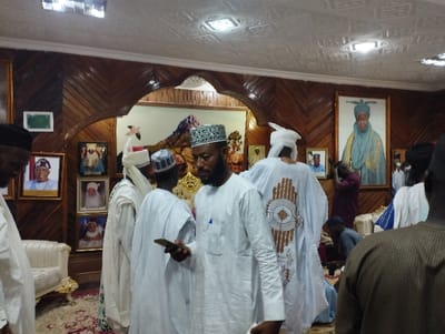 The image shows a group of men in a room in Kaltungo, Nigeria (latitude: 9.816, longitude: 11.315). The room is decorated with wood paneling on the walls and has a white ceiling with recessed lighting. There are several framed pictures on the walls, most of which depict men in traditional attire. The room has a white couch in the foreground, and a man in a white robe with brown embroidery is standing in the center of the image.  He is looking at a smartphone in his right hand, and holding a small white object in his left hand. Behind him is another man in a white robe with a white cap. On the right side of the image, another man in a white robe and a white cap is standing near a man in a brown and white striped shirt. The room appears to be a gathering space, possibly a community center or a meeting room.