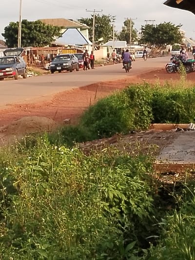 The image depicts a street scene in Agwala, Nigeria. A red dirt road runs diagonally across the frame, with a paved section to the right. Several vehicles are present, including a blue sedan on the left, a black sedan parked further down the road, and several motorcycles, some with riders. In the background, there are buildings with corrugated metal and tile roofs, some appearing to be market stalls or small shops. Trees and vegetation line the sides of the road. A few people are visible walking and standing near the vehicles and buildings. The sky is overcast, suggesting it is daytime.