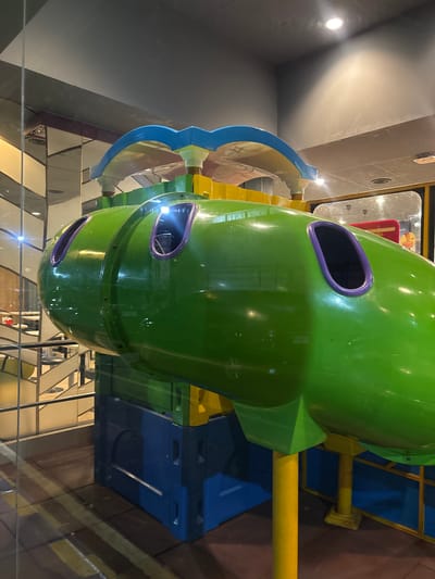 A green and purple spherical play structure is the prominent element in the image. It appears to be part of a children's playground. It is located within a building, as indicated by the ceiling and interior lighting. The structure is supported by yellow and blue beams or supports. The surrounding environment includes what looks to be a glass barrier, suggesting a viewing area. Given the context of Bencongan Indah, Indonesia, it is likely that the image was captured within a commercial area, potentially a family-friendly restaurant or mall, equipped with children's recreational facilities. There are no people visible in the image.