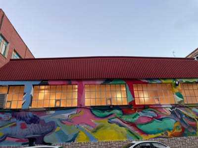 This media file shows a building with a large, colorful mural painted on its lower half. The building has a red, corrugated metal roof and large windows that are lit from within, casting a warm, orange glow. The windows are divided into many small panes. The mural is abstract and features vibrant colors such as pink, green, yellow, blue, and red. The style of the mural is dynamic and energetic, with swirling shapes and bold lines.
On the far left of the mural, there is a tag that reads "PAULA FRAILE 2026". The top portion of the image shows a clear, pale blue sky, suggesting it is either early morning or late evening. The lighting on the building and the sky indicate a transitionary period of the day. In the foreground, the top of a white car is visible, and the building's lower wall is made of textured stone blocks. The scene appears to be an urban or semi-urban environment, possibly an industrial building or a repurposed space that has been enlivened by street art. The overall impression is one of urban revitalization and artistic expression.