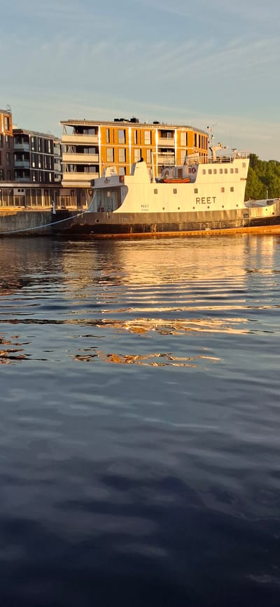 This image captures a serene waterfront scene in Pärnu, Estonia, likely during the late afternoon or early evening given the warm, golden light reflecting on the water and buildings. The primary subject is a white ferry boat, labeled with the name "REET" in large, black lettering on its side. The ferry has an orange section with multiple windows, suggesting it carries passengers or cargo. Adjacent to the ferry, modern buildings with numerous windows line the waterfront, their facades catching the sunlight. The water in the foreground is rippled, creating abstract patterns of light and shadow, with reflections of the ferry and buildings distorted by the movement. The sky is a clear, pale blue, indicating fair weather. The overall mood is peaceful and tranquil, highlighting the interplay of light, architecture, and water. Visible text includes "REET" on the ferry multiple times and "REET" on a smaller part of the ferry closer to the viewer.