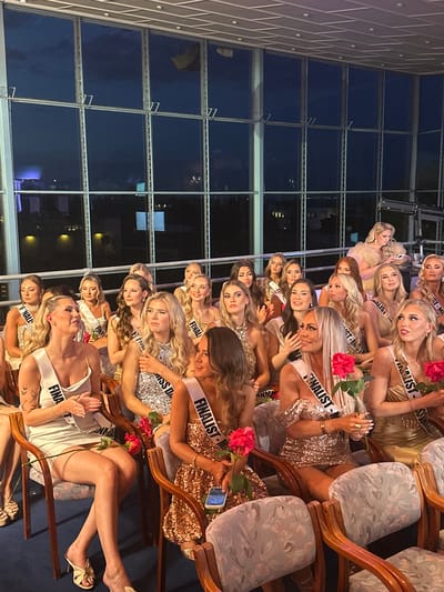 The image shows a group of women, likely beauty pageant contestants, wearing sashes that say "FINALIST" and variations thereof, such as "FINALIST-MISS," as well as roses. They are sitting in upholstered chairs with wooden frames. The backdrop is a large window offering a night-time view of what appears to be a city or urban area, and is in Skovshoved, Denmark. The women are wearing various dresses, some sequined. They appear to be in the midst of a beauty pageant event.