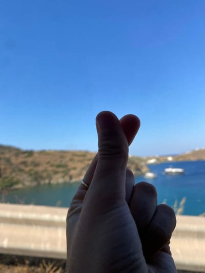 The image displays a human hand in the foreground forming a finger heart gesture, where the thumb and index finger touch to create a small heart shape. A thin, light-colored ring is visible on the ring finger of the hand. The hand is silhouetted against a brightly lit background.
The scene depicts a coastal environment in Greece, consistent with areas such as Φάρος (Pharos), characterized by a clear, bright blue sky occupying the upper portion of the frame. Below the sky, a large body of deep blue water, likely a bay or sea, stretches across the mid-ground. Arid, light-brown hillsides with sparse vegetation define the coastline on the left side of the frame. Several white vessels, appearing as boats or yachts, are scattered across the water, particularly towards the right side. In the far distance, indistinct light-colored structures are visible along the distant coastline, suggesting a settlement or collection of buildings. A light-colored barrier or railing extends horizontally across the lower mid-ground, indicating an elevated viewing position.