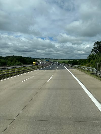 The image shows a view of the Autobahn A4 near Klipphausen, Germany. The highway is divided into multiple lanes, with traffic moving in both directions. In the foreground, the concrete road surface is clearly visible, with white lane markings. In the middle ground, several trucks and cars are traveling on the highway. To the left, a bright yellow truck is prominently featured. The surrounding landscape consists of green hills covered with trees and fields. The sky is overcast with dramatic, gray clouds. A guardrail runs along the side of the highway.