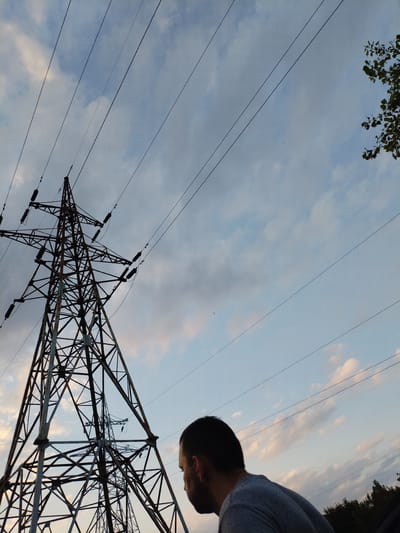 The image shows a low-angle shot of a large electricity pylon against a sky with clouds. A man is visible in the foreground, looking up and to the right, towards the pylon. The sky is a mix of blue and gray clouds, suggesting either early morning or late afternoon. Several power lines stretch across the frame, originating from the pylon. In the background, another, smaller pylon can be seen, and at the far right, the tops of trees are visible. The overall impression is one of an outdoor industrial or semi-rural setting. No visible text is present in the image.