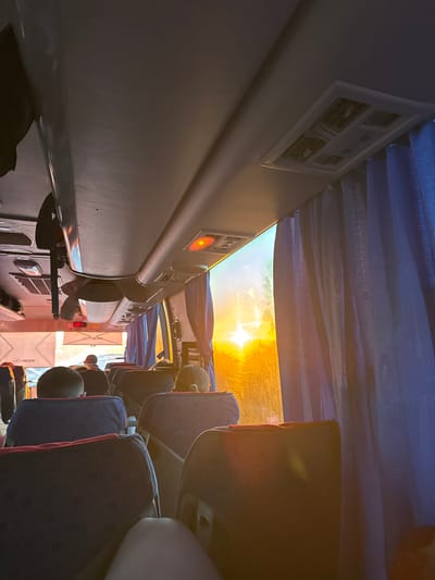 The image depicts the interior of a bus. The viewer is positioned in an aisle seat, looking toward the front of the bus, as the image has been rotated. Seats upholstered in dark blue with red trim are visible on the right side of the aisle. Passengers' heads are visible between the seats. On the left side, the wall of the bus includes vents, lights, and an emergency exit window. Bright sunlight streams through the front window, partially obscured by blue curtains. The brand name "Higer" is displayed on a placard on the front of the bus. The location, Ustinovo, Russia, is not explicitly supported by elements in the image.