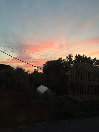 The image depicts a rural scene at dusk in the village of Maloye Verevo, Russia. The sky is painted with soft hues of pink and orange from the setting sun, with scattered clouds. Silhouetted against the sky are the dark shapes of trees and overhead power lines. In the foreground, a partially constructed wooden house frame stands prominently on the right side of the image. To the left, a dark, leafy canopy obscures some of the scene, with the roof of a dark-colored building visible in the upper left corner. Further left and slightly lower, a silver, dome-shaped structure, possibly a greenhouse or storage unit, is visible amongst the vegetation, behind a dark wooden fence. The ground in the foreground is covered with dark gravel.