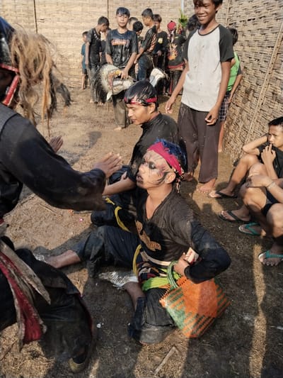 This image captures a group of young men and boys outdoors in Kedawung, Indonesia, seemingly during a break or after a traditional performance. The ground is muddy, indicating a physically demanding activity involving dirt and water.
In the foreground, a man with long, dark, unkempt hair and black clothing is partially visible on the left. His arm is extended towards a central figure, a young man seated on the muddy ground. This central figure, likely a performer, wears a black long-sleeved shirt, black pants, and a red bandana with streaks of blue hair. His face is elaborately painted with black, white, and red, resembling a tiger or mythical creature. He holds a brightly woven orange and green plastic basket. His expression suggests weariness or a state of trance.
Around these central figures, several other young men and boys are present. One boy in a white and black t-shirt stands smiling broadly in the background. Another boy leans against a woven bamboo wall, wearing a green sleeveless shirt and checkered shorts. Several others are holding traditional masks or headdresses, also covered in mud, suggesting they are props from the performance. These masks often represent mythical animals and are characteristic of Indonesian traditional trance dances such as Kuda Lumping or Jathilan.
The setting is outdoors, with a distinct woven bamboo wall forming the backdrop, a common feature in rural Indonesian environments. The bright daylight and visible shadows indicate it is daytime, likely late morning or afternoon. The participants are largely covered in mud, highlighting the immersive and physical nature of the event. While some appear tired, others seem relaxed and observing. Visible text on the face-painted man's shirt includes partial words like "GEMB" and "UNJ", and on a green shirt, "HANG ANJ". The scene offers a raw, authentic glimpse into a local cultural tradition.