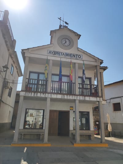 This is a full-frame, eye-level, outdoor, daytime, medium shot of the Cadalso, Spain Town Hall (Ayuntamiento). The building is two stories tall with a central clock tower on its roof. The clock face shows the time as approximately 10:12. There are several flags displayed on the second-story balcony: a green and yellow flag, a Spanish flag, and a black and white flag. The building's facade is white with large pillars supporting a covered walkway on the ground floor. There is a window and a wooden door on the ground floor, with a bench placed in front of the window. The surrounding environment consists of narrow streets and other buildings. The sky is clear and blue, with the sun shining brightly and casting shadows.