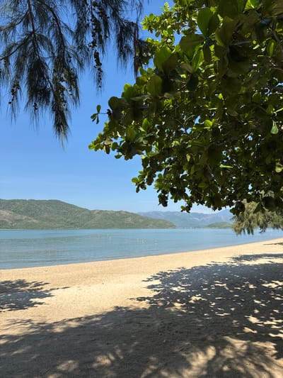 The image shows a serene beach scene in Hoa Thang, Vietnam. The foreground is dominated by a sandy beach with dappled sunlight and shadows from overhanging trees. In the midground, calm, blue water meets the shore, with a line of orange buoys visible in the distance. The background features rolling green hills and mountains under a clear blue sky. The overall impression is one of a peaceful, tropical paradise. There are no people or animals visible in the frame. The foliage in the foreground suggests the presence of lush vegetation typical of a coastal environment in Vietnam.