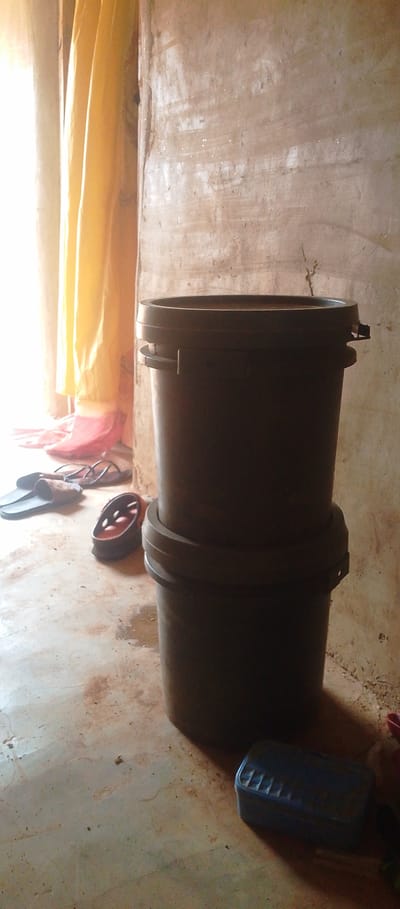 This is a medium-shot, natural light photograph taken indoors, likely in a home setting in Achara Ndiagu Omegio, Nigeria. The scene depicts two stacked, dark-colored plastic buckets in the foreground, set against a roughly textured, light brown wall. Sunlight streams in from the left, illuminating yellow curtains and casting a bright glow on the wall. On the floor to the left of the buckets are a pair of brown flip-flops and a pair of black sandals. A dark red object, possibly a plastic container, is visible behind the sandals. A blue plastic container with a ridged lid rests on the floor in the lower right corner of the image, partially in shadow. The floor itself appears to be a concrete or dirt surface, with some dirt and staining visible. The overall impression is of a humble, everyday domestic environment. The time of day is likely morning or afternoon, given the strong sunlight. There is no visible text in the image.