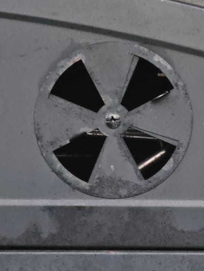 A close-up, slightly angled, eye-level shot shows a circular vent with four triangular openings on a dark gray, textured surface. The vent is made of metal and has a rusted, weathered appearance. A single screw is visible in the center of the vent. The surface appears to be part of an appliance or piece of equipment. The environment is not visible, and no location in Mäntsälä, Finland, can be determined from the image.