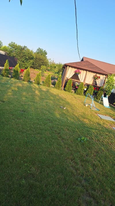 The image depicts a sunlit backyard in the city of Sărata-Galbenă, Moldova. A large expanse of manicured green grass dominates the foreground, sloping gently upwards towards a raised garden bed lined with small, conical evergreen trees and red flowers. In the middle ground, a brown-roofed gazebo with cream-colored curtains is visible, adorned with hanging flower baskets. Adjacent to the gazebo, a small blue and white soccer goal with a ball, and a white children's slide are set up on the lawn. A dark, thin cable hangs diagonally across the top right of the frame against a clear blue sky. Further back, a brown wooden fence separates the property from a wooded area with lush green trees. The overall scene suggests a well-maintained outdoor recreational space.