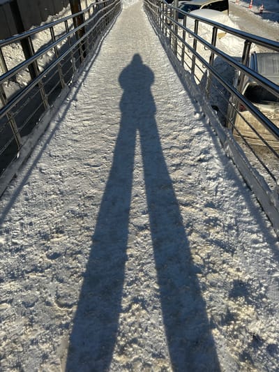The image depicts a snow-covered pedestrian ramp or bridge under clear, low-angle sunlight, casting a prominent shadow of a person. A long, distinct human shadow, likely belonging to the photographer and indicating a hooded figure, stretches from the bottom center of the frame along the snow-covered walkway towards the upper center. The walkway surface is uneven, consisting of compacted snow and ice, with visible textured patterns and faint footprints. Polished metal railings, appearing to be stainless steel with multiple horizontal bars, line both sides of the pathway, extending into the background. To the left of the walkway, beyond the railing, is a snow-covered embankment or raised concrete structure. To the right, at a lower elevation, a street or parking area is visible with several vehicles, including a light-colored sedan partially obscured by snow, and orange traffic cones in the distance. The overall environment suggests winter conditions in an urban or suburban setting. Specific street or landmark identification within Shul'gino, Russia, is not discernible from the visual information provided.