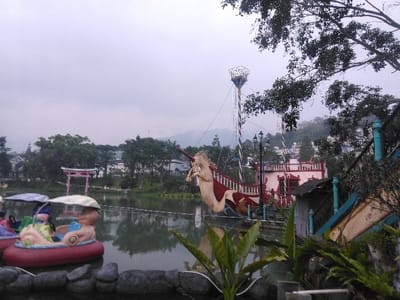 The image depicts an outdoor amusement park scene within the Parque de Diversiones Dr. Roberto Ortiz Brenes in Ciudad de San José, Costa Rica, under an overcast sky.
In the left foreground, a dark, low barrier separates the viewer from a body of water. Partially visible are two brightly colored paddle boats, or similar watercraft, docked or stationary in the water. The primary visible boat is red with a light-colored, cartoonish character design forming its upper half, featuring a smaller figure seated inside.
The middle ground is dominated by a calm lake, reflecting the gray sky and distant foliage. Across the lake, a prominent pink Japanese-style torii gate stands centered towards the left.
To the right of the torii gate and extending over the water is a large, elaborate amusement ride featuring a stylized dragon or sea serpent with a horse-like head, colored cream, red, and brown. This structure connects to a tall, vertically striped pole (blue and white) topped with a cage-like observation platform or mechanism. An ornate black streetlamp with multiple globes is positioned near the base of this pole. Behind the dragon ride and pole, a white building with red railings is visible on a sloping terrain.
The background features numerous green trees and several residential-style buildings with light-colored walls and red roofs, situated on distant hills under the overcast sky. A section of a light-colored slide or ramp with blue railings is visible on the far right, partially obscured by dense green foliage, which also extends into the upper right corner of the frame. No individuals are clearly identifiable within the scene.