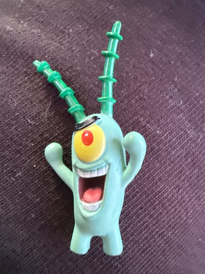 The image shows a close-up, overhead view of a light green plastic toy figure of Plankton from SpongeBob SquarePants. The toy is lying on a dark, textured surface, possibly fabric. Plankton is depicted with its characteristic wide mouth open, showing pink inside and white teeth. It has a large yellow eye with a red pupil and a black band across it. Two segmented green antennae extend from the top of its head. Its arms are raised. The background is a dark, woven material with visible threads and some small white specks. There is no indication of the city of Vyzyrka, Ukraine in this image.