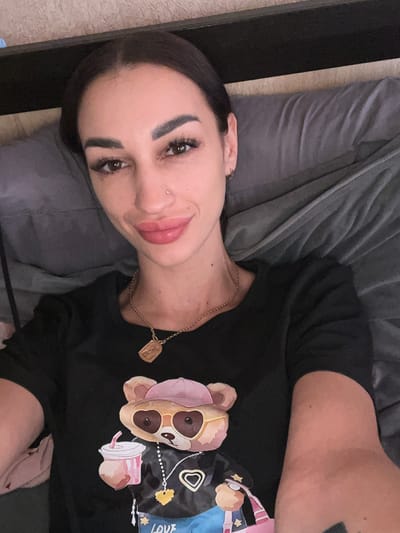 The image shows a woman lying on a bed, looking at the camera. She has dark hair pulled back, dark eyebrows, and is wearing makeup. She has a nose ring and hoop earrings. She is wearing a black t-shirt with a graphic of a cartoon bear holding a drink. She is wearing a gold necklace with a rectangular pendant. The background appears to be a bedroom, with a textured wall and a gray blanket. The image was taken in Bashtanka, Ukraine.