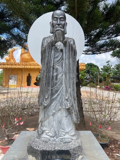 A tall, dark grey stone statue of an elderly male figure with a long beard and flowing robes stands centrally in an outdoor setting in Đăk Mar, Vietnam. The figure's hands are clasped at chest level, and a large, circular, white disc is positioned directly behind its head, resembling a halo. The statue rests on a dark stone base with Vietnamese text inscribed. To the left, a golden multi-tiered architectural structure, likely a temple, is visible, featuring smaller statues within its archways. Sparse, potted deciduous trees with some red/pink blossoms flank the main statue in the foreground. The ground consists of bare earth and patches of green foliage with pink flowering plants in the midground to the right. The background features more trees and utility poles under a partly cloudy sky. No people are visible.