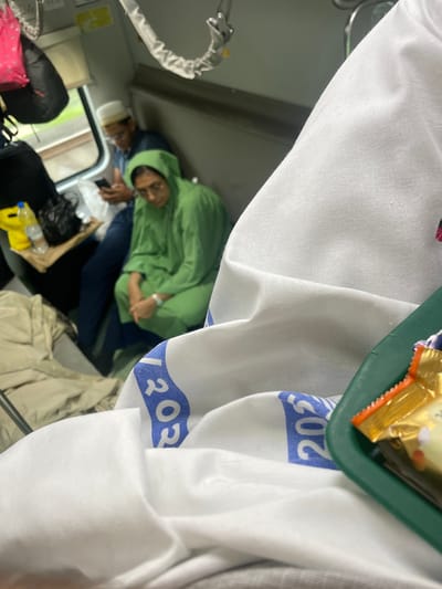 The image depicts the interior of a train carriage, likely in Dahanu, India, given the context. In the foreground, a white cloth with blue markings is draped over a surface, partially obscuring a green tray containing candy wrappers. In the middle ground, a woman wearing a bright green headscarf and garment is seated, looking downwards. Beside her, a man wearing a white cap and glasses is holding a mobile phone. The background shows luggage, including a pink bag and a black backpack, and a window offering a view of the outdoors. The lighting suggests it is daytime.