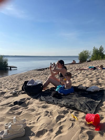 The image depicts a beach scene in Matiushino, Russia. A woman in a black bikini and black cap is seated on a dark blanket with a child. The child is eating and is covered by a blue towel. The woman is holding a mobile phone. Beside them is a black bag, along with white sandals and other footwear in the sand. In the background, there is a body of water, trees, and other people sunbathing. A red bucket and a yellow shovel are visible on the left side of the blanket.