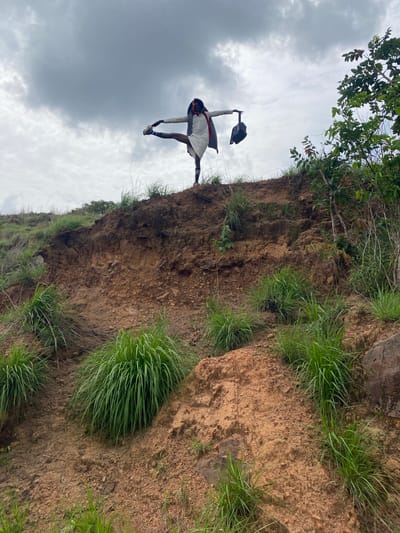 The image, taken from a low angle, shows a person standing on the edge of a dirt cliff or embankment. The person is dark-skinned, wearing a white tunic with black sleeves, socks, and shoes. The person's arms are outstretched and one leg extended, with the person's hand grasping their foot. They also hold a black object, possibly a bag, aloft in one hand.
The cliff is composed of brown earth and sparse green grass, and it rises above the viewer. A tree with green leaves is partially visible in the top left corner of the image. The sky is overcast with gray clouds.
While the prompt mentions Amaigbo, Nigeria, this information is not evident from the visual information provided in the image itself. Therefore, Amaigbo is not a conclusion that can be drawn from the photograph.